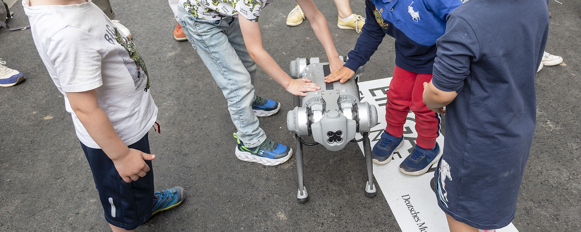 Fünf Kinder stehen um einen grauen vierbeinigen Roboter auf Asphalt; zwei Jungen berühren den Roboter, einer trägt rote Hose und blauen Pullover, ein anderer hellblondes Kind links beobachtet, im Hintergrund Erwachsene und ein Schild mit teilweise sichtbarem Text "Deutsches Museum".