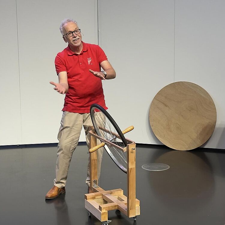 Ein Mann in rotem Poloshirt und beiger Hose steht in einem Vortragsraum des Deutschen Museums und demonstriert ein Holzgestell mit einem montierten Fahrrad-Vorderrad.