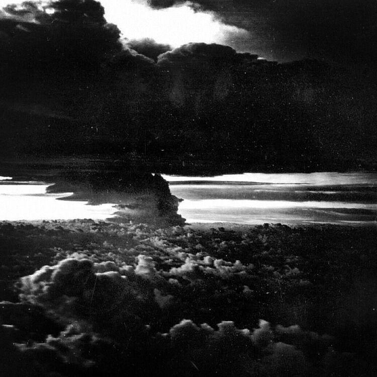 Schwarzweiß-Foto einer dramatischen, hoch aufragenden Pilzwolke über einer Schicht aus dichten, aufgewölbten Wolken, mit hellem Horizontlicht und dunklem, stürmischem Himmel darüber.
