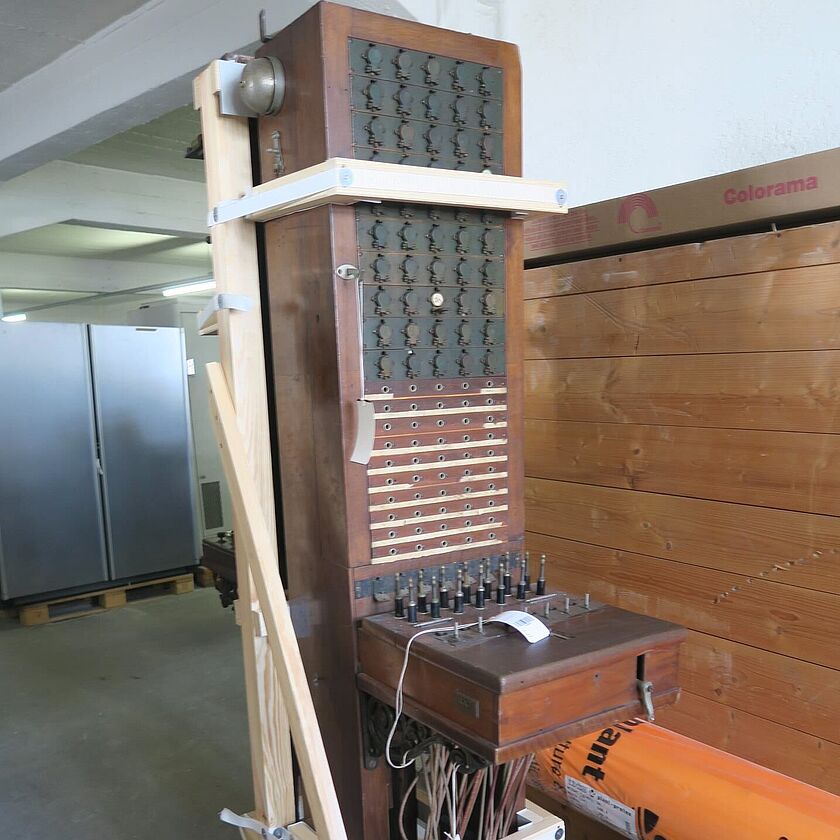 Quaderförmiger, aufrechtstehender Holzkasten mit Buchsen und von unten hingeführten Verbindungskabeln (Klappenschrank). Gehalten durch Holzgerüst im Depot.