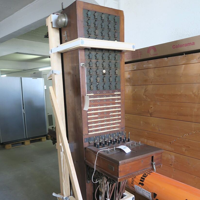 Quaderförmiger, aufrechtstehender Holzkasten mit Buchsen und von unten hingeführten Verbindungskabeln (Klappenschrank). Gehalten durch Holzgerüst im Depot.