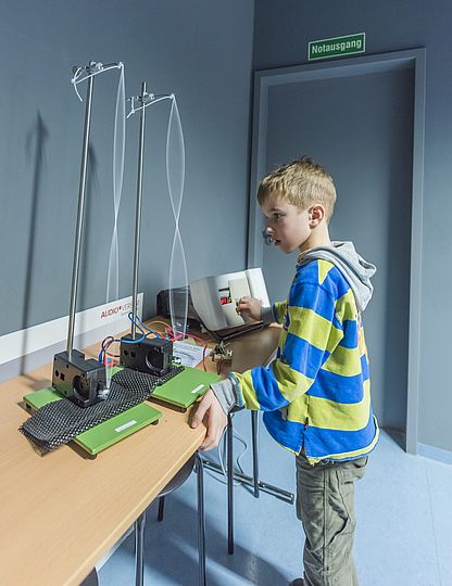 Ein Junge schaut sich Schwingungen von zwei vertikal eingespannter Seilen an. Diese zeigen die Schwingungen eines Tons an. Der Ton wird von einem Gerät aus auf die beiden Seile übertragen.