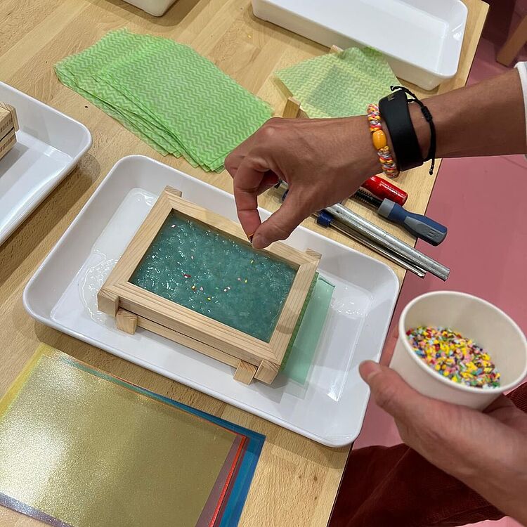 Person steckt bunte Streusel in eine rechteckige Holzform mit blau-grünem Papierbrei auf einem weißen Tablett, auf dem Tisch liegen bunte Pergamentbögen, grüne Tücher und Werkzeuge; rechter Hand hält die Person einen Pappbecher mit Streuseln.