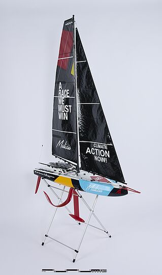 Modell eines Rennseglers auf Ständer mit schwarzem Großsegel, Schriftzug „A RACE WE MUST WIN“ links und „CLIMATE ACTION NOW!“ rechts, bunte Rumpfbemalung und roter Kiel.