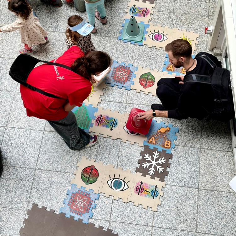 Auf dem Boden liegt ein großes Brettspiel aus weichen Puzzle-Matten. Auf den Feldern sind verschiedene Symbole wie eine Schneeflocke, ein Auge oder ein Atom aufgedruckt. Ein Mann, eine Museumsmitarbeiterin im roten T-Shirt und einige Kinder spielen gemeinsam, wobei der Mann gerade eine große rote Spielfigur bewegt.