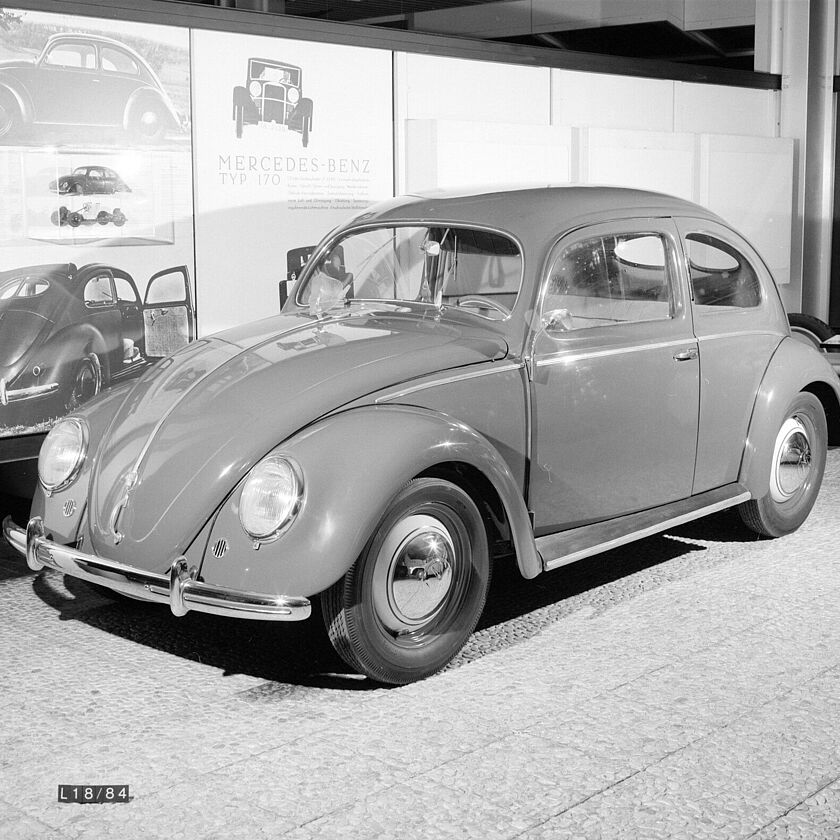 2025_03_2025-1 Schwarzweiß Aufnahme eines VW Käfers in den Ausstellungsräumen vor bebilderten und beschrifteten Ausstellungswänden. Das Auto ist von vorne rechts aufgenommen. Am Bildrand unten Links ist ein schwarzes Täfelchen mit der Aufschrift "L18/84", vermutlich die Inventarnummer des Käfers, zu erkennen.