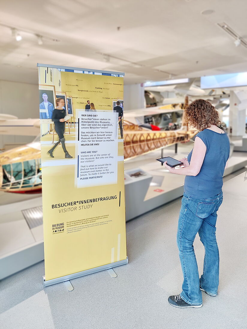 Teilnehmerin beim Ausfüllen der Umfrage zur Leibniz-Besuchendenstrukturanalyse vor einem gelben Banner in der Ausstellung Luftfahrt