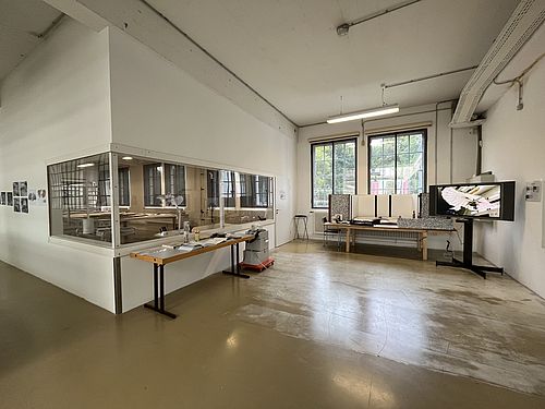 Das Atelier in der Flugwerft Schleißheim Ein Raum mit Fenstern nach innen und Fenstern nach außen. Davor stehen Vitirinen und ein Bildschirm.