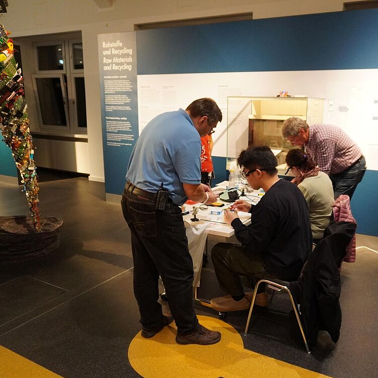 Fünf Personen an einem Workshop-Tisch in einer Museumsausstellung zum Thema Rohstoffe und Recycling: zwei Männer stehen, drei Teilnehmende sitzen und arbeiten an kleinen Elektronik- oder Bastelteilen; im linken Bildrand eine hohe, schrauben- und platinenbestandene Skulptur; Informationstafeln und Fenster im Hintergrund.