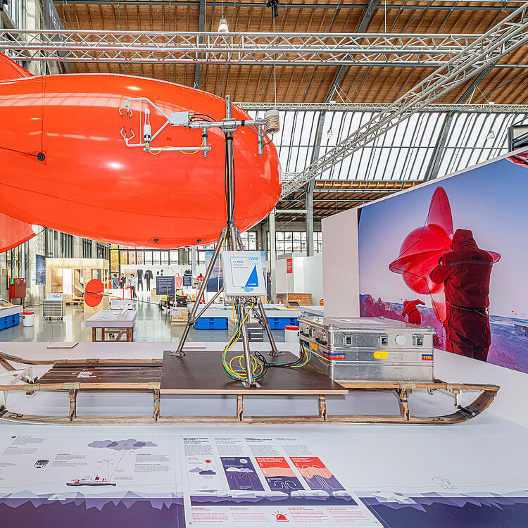 Ausstellungsexponat der Sonderausstellung 'Dünnes Eis': Ein roter Heliumballon namens 'Miss Piggy' aus der Expedition POLARSTERN. Der Ballon ist mit einer Wetterstation und Messgeräten auf einem Schlitten befestigt. Im Hintergrund sind Ausstellungstafeln, technische Details und eine Fotografie eines Forschers mit dem Ballon in der Polarregion zu sehen.