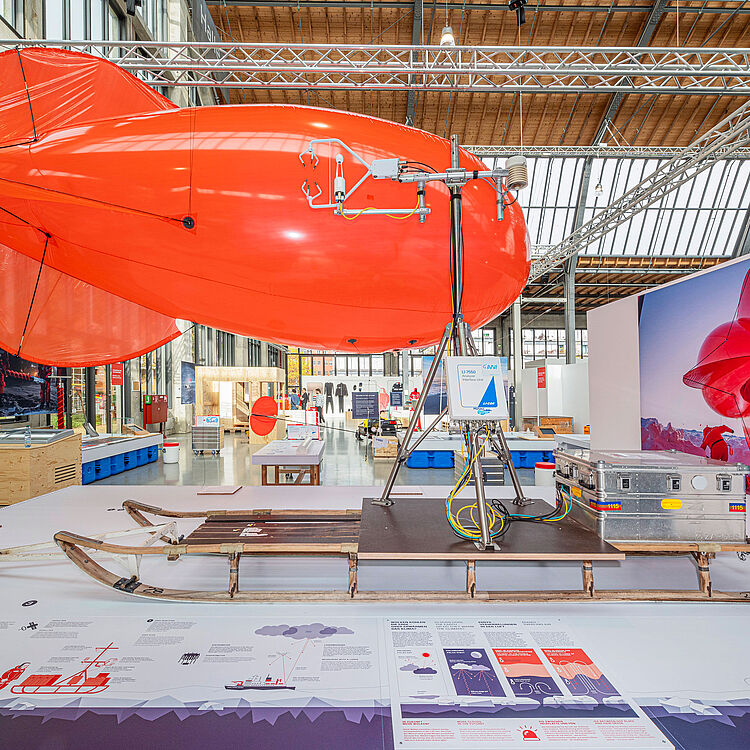 Ausstellungsexponat der Sonderausstellung 'Dünnes Eis': Ein roter Heliumballon namens 'Miss Piggy' aus der Expedition POLARSTERN. Der Ballon ist mit einer Wetterstation und Messgeräten auf einem Schlitten befestigt. Im Hintergrund sind Ausstellungstafeln, technische Details und eine Fotografie eines Forschers mit dem Ballon in der Polarregion zu sehen.