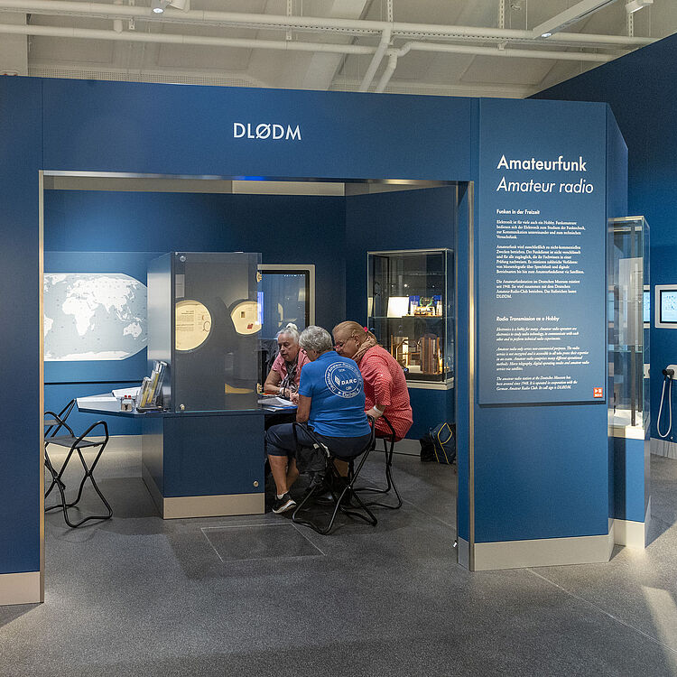 Drei Funkamateure sitzen an einem Tisch vor der Funkstation DLØDM und unterhalten sich. Über ihnen hängt ein Schild mit der Aufschrift "Amateurfunk – Amateur Radio" und dem Rufzeichen DLØDM. Im Hintergrund befindet sich eine Weltkarte, die Funkreichweiten darstellt. In den Vitrinen sind historische Geräte ausgestellt.