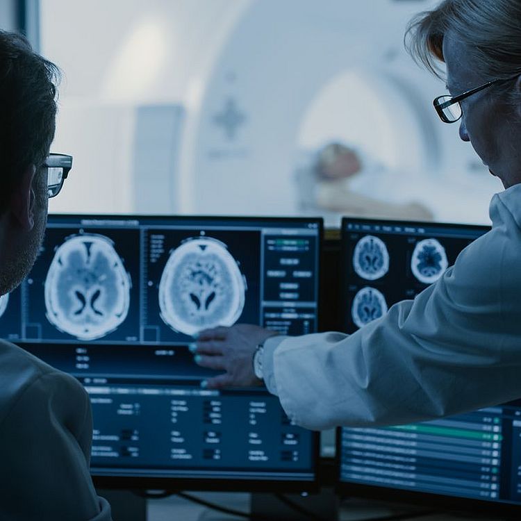 Im Kontrollraum besprechen Arzt und Radiologe die Diagnose, während sie das Verfahren und die Monitore mit den Ergebnissen der Gehirnscans beobachten, während der Patient im Hintergrund eine MRT- oder CT-Untersuchung durchführt.