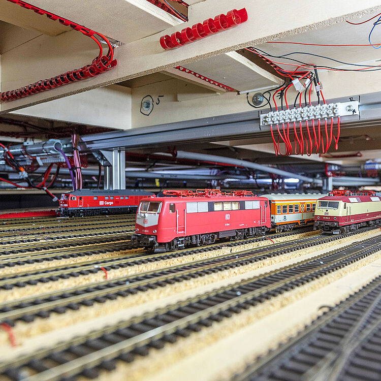 Die Modellbahn des Deutschen Museums hinter den Kulissen. Mit mehreren Gleisen und Modellzügen, darunter zwei auffällige Lokomotiven in Rot und Beige. Die Unterkonstruktion zeigt eine komplexe Verkabelung für die Steuerung der Anlage.