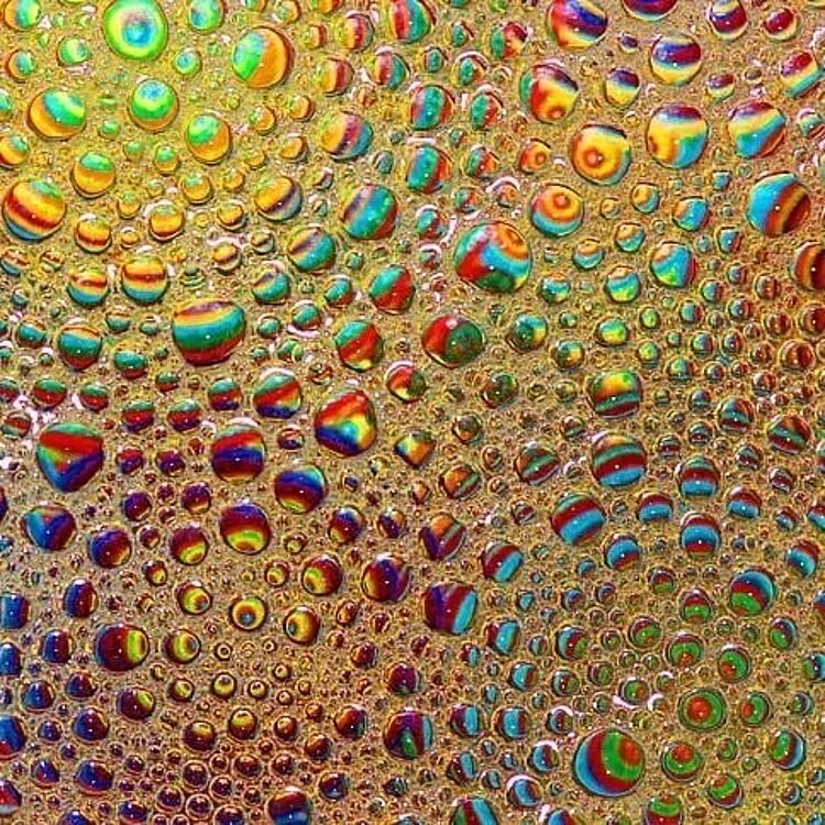 Bunte, glänzende Wassertropfen in unterschiedlichen Größen auf einer Oberfläche, die durch Regenbogenreflexe lebhafte Streifen in Rot, Blau, Grün und Gelb zeigt; enge Tropfenmuster bilden ein texturiertes, fast psychedelisches Mosaik.
