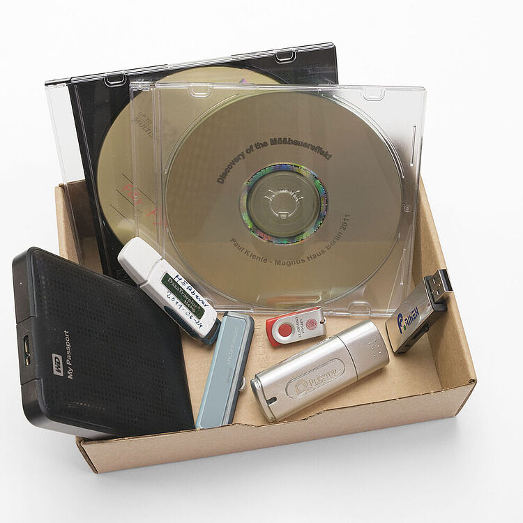 Eine flache Pappschachtel mit mehreren Speichermedien: zwei CD/DVD in durchsichtigen Hüllen, ein schwarzes externes USB-Festplattengehäuse, vier verschiedene USB-Sticks und ein schmaler blauer SD-/USB-Adapter.