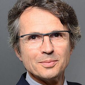 Porträtaufnahme einer erwachsenen Person mit schulterlangem, leicht gewelltem graumeliertem Haar, ansprechendem dunklen Sakko und weißem Hemdkragen vor neutralem grauem Hintergrund; das Gesicht ist unkenntlich gemacht.