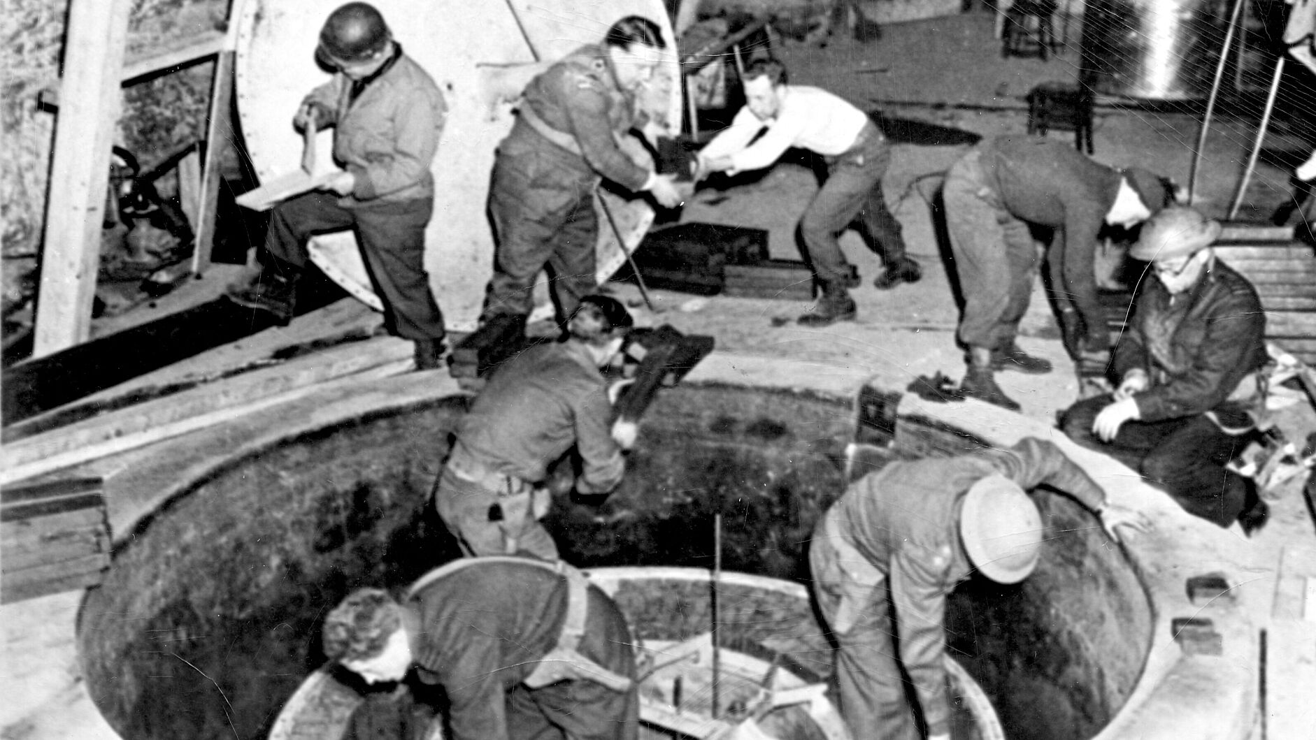 Fotografie. Acht Männer in Uniform, teils mit Helm, verrichten Arbeiten an einem im Boden eingelassen ca. 2 Meter breiten Apparat.