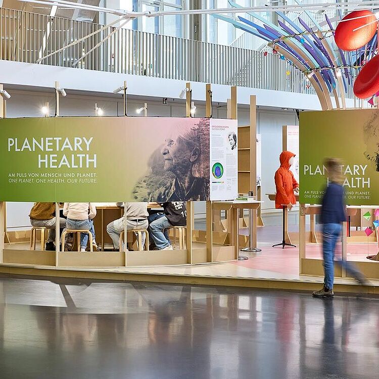 Sonderausstellung mit großem grünen Banner „PLANETARY HEALTH“, rundem Holzrahmen und Besuchern auf Hockern; rechts bunte Skulptur, rote Jacke auf Kleiderständer und eine verschwommene Person, die vorbeigeht.