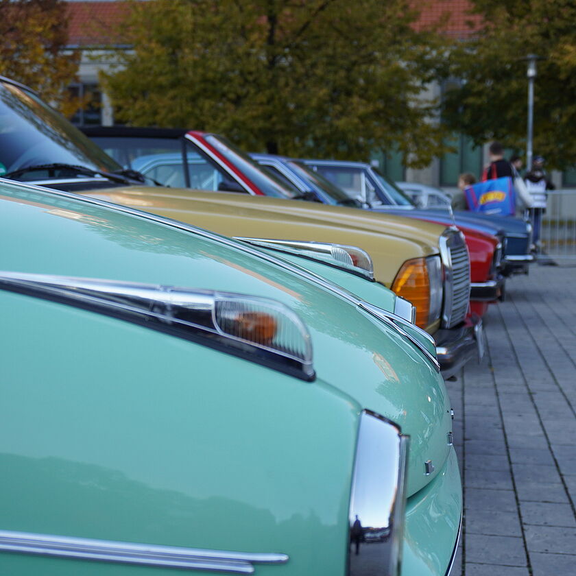 Eine Reihe farbenfroher Oldtimer auf dem Vorplatz des Verkehrszentrums, im Vordergrund ein mintgrünes Auto mit Chromdetails, dahinter weitere klassische Fahrzeuge in Gelb, Rot und Blau.