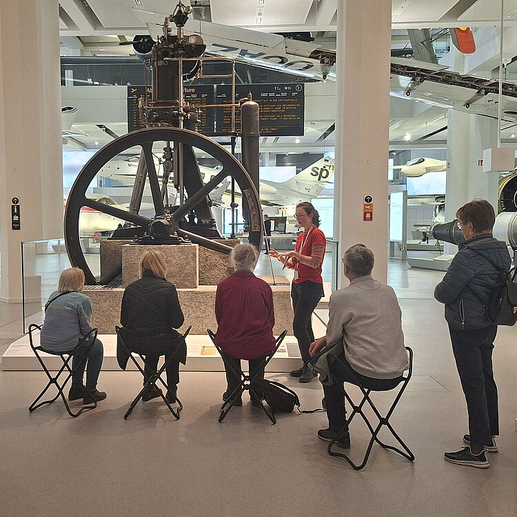 Eine Besuchergruppe in der Ausstellung "Energie – Motorem". Ein Museumsguide erklärt vor einer großen historischen Dampfmaschine mit großem Schwungrad, mehrere Personen sitzen auf kleinen Klappstühlen.