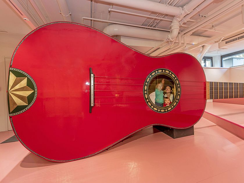 Drei Kinder spielen mit der Riesengitarre. Zwei von ihnen sind in der Gitarre, ein Junge zupft von außen eine Seite.