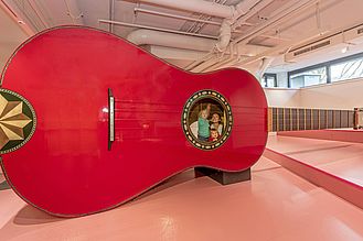 Three children playing with the giant guitar. Two of them are inside the guitar, one boy is plucking a string from the outside.