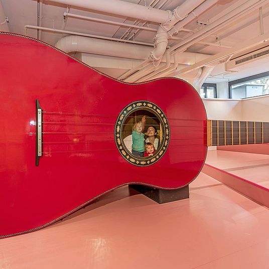 Drei Kinder spielen mit der Riesengitarre. Zwei von ihnen sind in der Gitarre, ein Junge zupft von außen eine Seite.