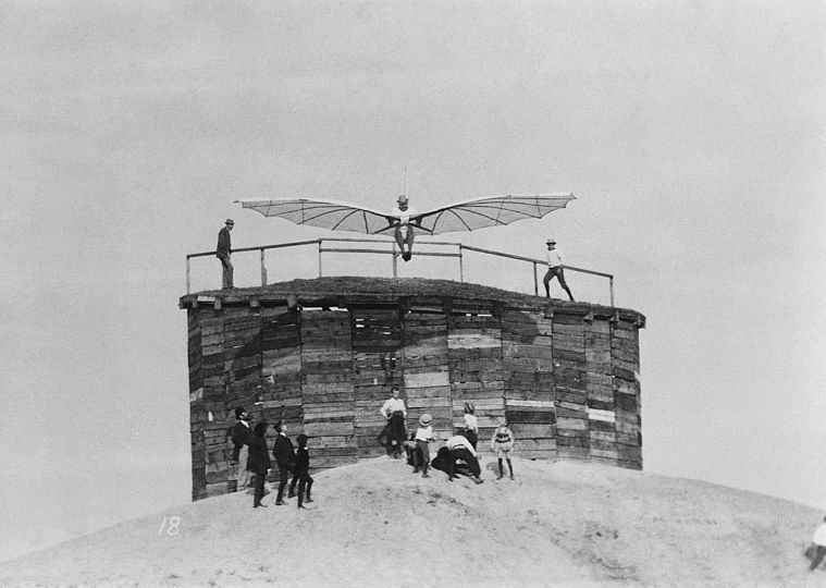 Otto Lilienthal steht mit seinem Segelapparat auf einer Konstruktion, um ihn herum Zuschauer, er ist kurz vor dem Start. 