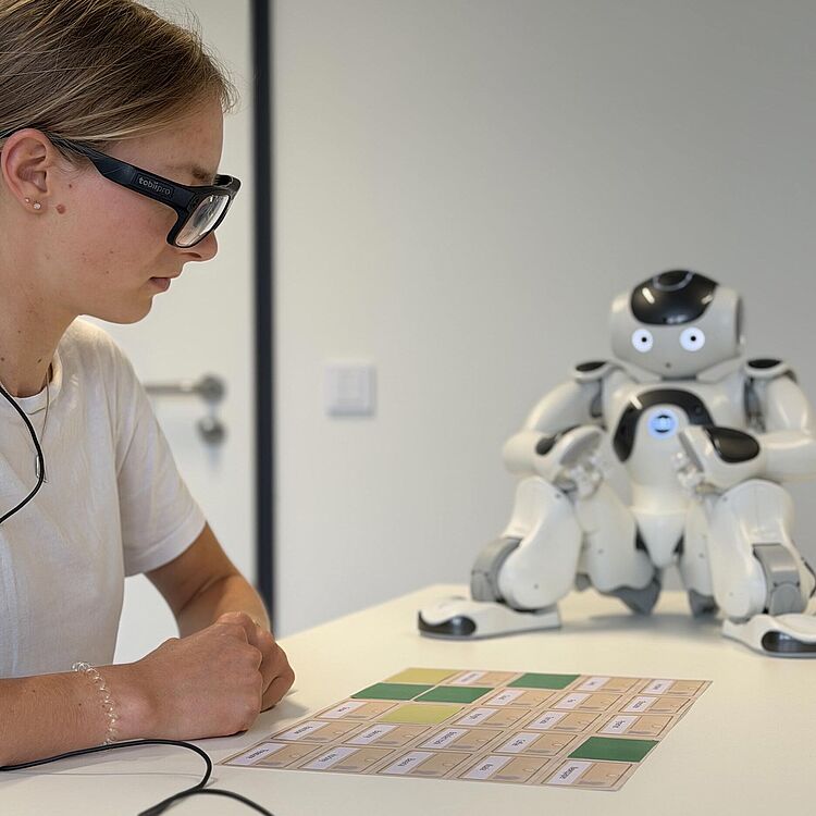 Nahaufnahme einer Person mit Eye-Tracking-Brille, die an einem Tisch sitzt und auf ein beschriftetes Kartenraster blickt, gegenüber ein kleiner weißer humanoider Roboter NAO auf dem Tisch.