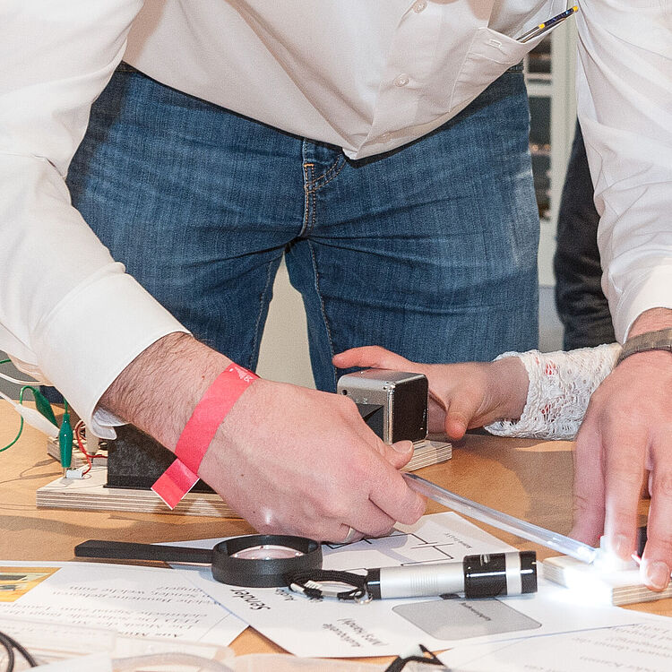 Ein Erwachsener und ein Mädchen beugen sich über einen Holztisch und montieren ein kleines Elektronik-Projekt mit LED-Streifen, Drähten, Krokodilklemmen, einem Taschenlampen-ähnlichen Licht und Messwerkzeugen; beide tragen helle Oberteile, im Hintergrund ein Büro mit Regalen und weiteren Personen.