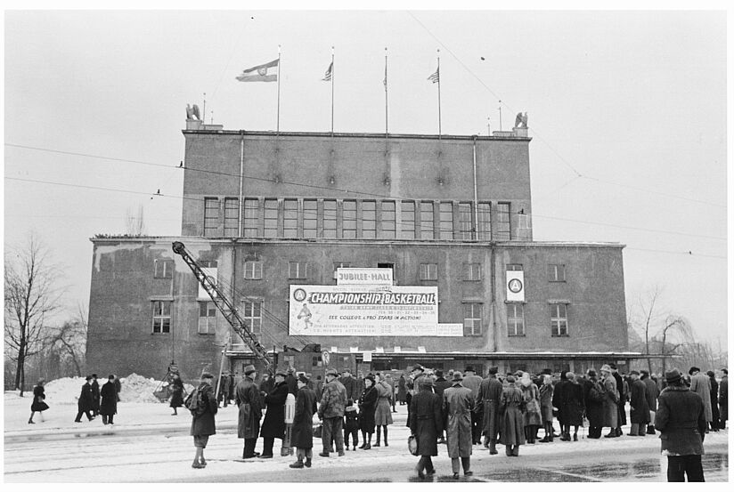 Blick auf den Kongresssaal, mit Fahnen beflaggt und Werbetafel, davor steht auf der Straße eine Menschenmenge.