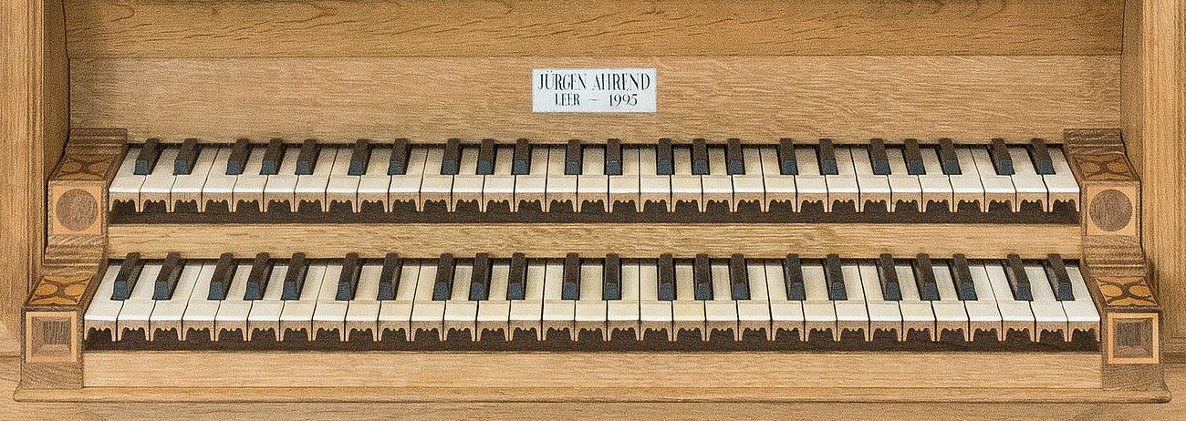 Zwei übereinanderliegenden Manualtastaturen der Ahrend-Orgel, mit einer Inschrift über den Manualen: „Jürgen Ahrend, Leer – 1995“. Die Tasten sind in klassischem Stil gehalten, mit weißen Untertasten und schwarzen Obertasten. Die Umrahmung der Tastaturen besteht aus hellem Holz mit kunstvollen Einlegearbeiten an den Seiten.