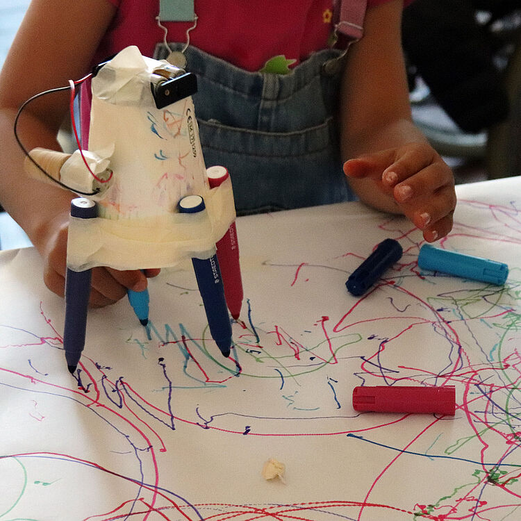 Ein Kind nimmt am Workshop "Malen mit Strom und Fliehkraft" teil und experimentiert mit einer selbstgebauten Malmaschine. Die Maschine, bestehend aus einem Papierbecher, Filzstiften und Klebeband, bewegt sich über ein großes Blatt Papier und hinterlässt bunte, zufällige Linien.