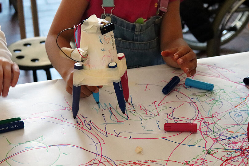 Ein Kind nimmt am Workshop "Malen mit Strom und Fliehkraft" teil und experimentiert mit einer selbstgebauten Malmaschine. Die Maschine, bestehend aus einem Papierbecher, Filzstiften und Klebeband, bewegt sich über ein großes Blatt Papier und hinterlässt bunte, zufällige Linien.