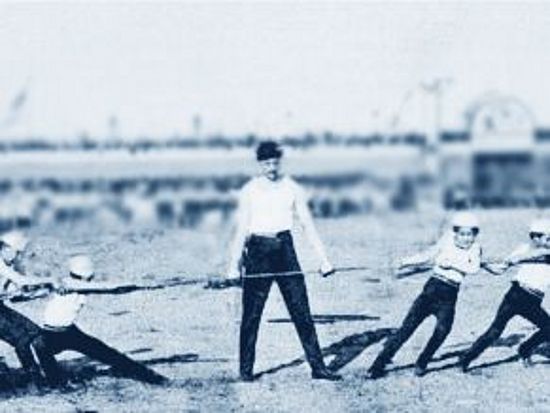 Kooperation und Konkurrenz Kinder ziehen Seil. Links ziehen 4 Kinder, am rechten Ende 5 Kinder. In der Mitte steht ein Mann. Er hält das Seil mit seiner linken Hand fest.