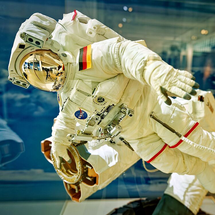 Ein weißer Raumanzug mit goldverspiegeltem Helmvisier und NASA-Logo, an einem Modell oder Schaufenster befestigt, im Schwebeflug-Posing; auf dem Arm ein deutsches Flaggenpatch, zahlreiche Verbindungskabel, Halterungen und rote Markierungen sichtbar, Hintergrund mit wolkenartiger Weltraum- oder Museumsdekoration.