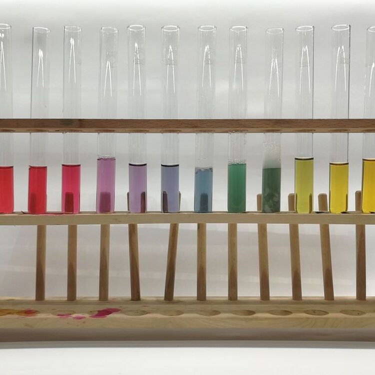 Das Bild zeigt eine Reihe von Reagenzgläsern in einem Holzständer, die Flüssigkeiten mit unterschiedlichen Farben enthalten. Diese Farben repräsentieren den pH-Wert einer Lösung, von Rot (sauer) über Lila (neutral) bis Gelb (basisch).