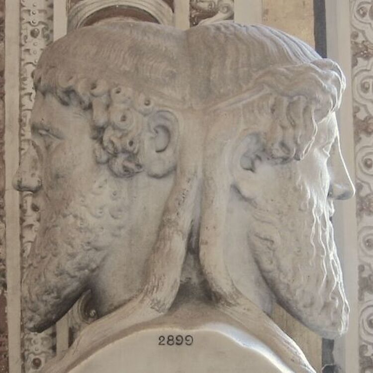 Marmorskulptur eines zweigeschichtigen Janus-Kopfes mit zwei gegensätzlichen, bärtigen Profilen, feinen Locken und nummerierter Basis (2899), vor ornamentaler Wand.