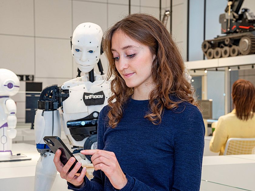 Junge Museumsbesucherin blickt auf ihr Smartfone, welches sie in der Hand hält. Im Hintergrund steht ein menschlich aussehender Roboter.