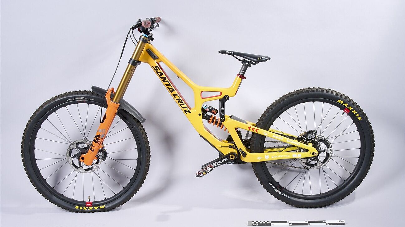 Mountainbike von der Seite fotografiert. Besonderheiten: gelber Rahmen, hohe Federung, große Reifen. Es ist deutlich, dass es sich um ein stabiles und optimiertes Sportgerät handelt.