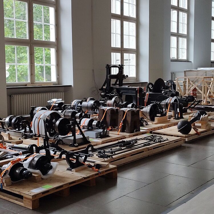 Große Halle mit hohen Fenstern, in der zahlreiche industrielle Metallteile, Zahnräder, Trommeln und Maschinenkomponenten sorgsam auf Holzpaletten und in Holzkisten verpackt und mit orangenen Spanngurten fixiert sind; heller Steinboden und große Fensterfront links, im Hintergrund massive hölzerne Pressteile.