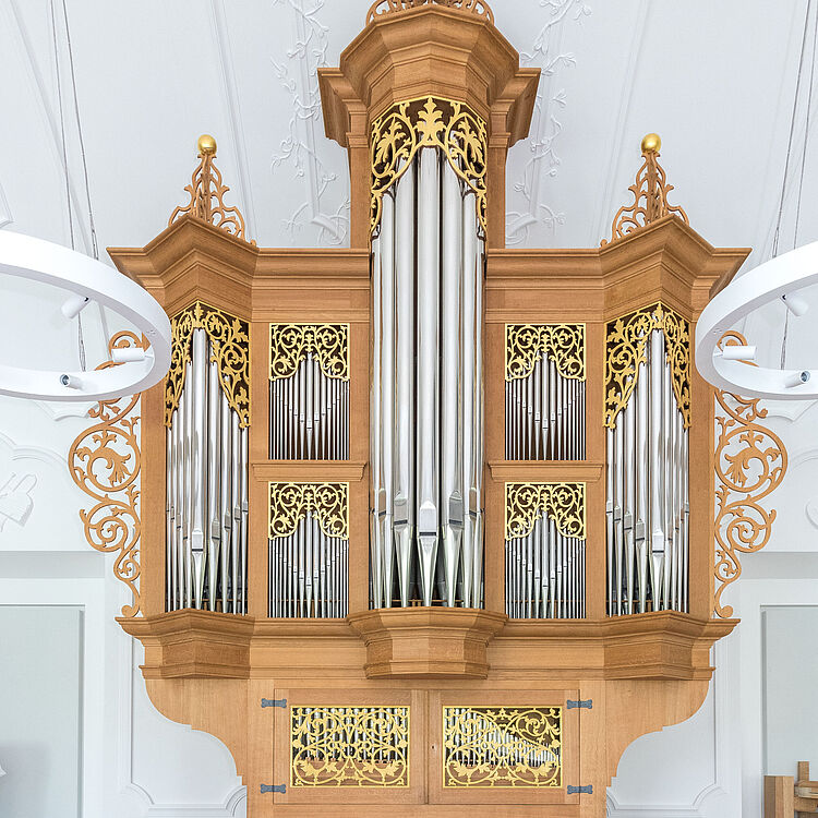 Das Bild zeigt die Ahrend-Orgel. Hierbei handelt es sich um einen Nachbau einer typisch norddeutschen Barock-Orgel, die eigens für das Deutsche Museum angefertigt wurde. Sie ist etwa 6 Meter hoch, besteht zum Großteil aus hellem Holz, mit geschwungenen Verzierungen an den Enden. Zu sehen ist der Spieltisch mit Klaviatur und Registerzügen. Darüber befindet sich der Aufbau der Pfeifen-Werke, wobei die größten Zungenpfeifen zentral angeordnet sind. 