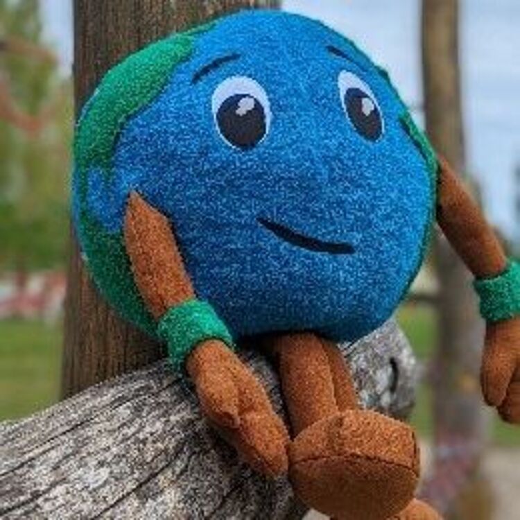 Plüschspielzeug in Form eines lächelnden Erdballs mit blauen Ozeanen, grünen Kontinenten, braunen Armen und Beinen, das auf einem verwitterten Holzgeländer vor unscharfem Outdoor-Hintergrund sitzt.