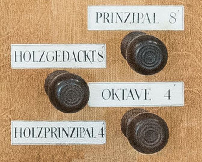 Vier runde Registerzüge der Ahrend-Orgel auf hellem Holzuntergrund. Die Beschriftungen lauten: Prinzipal 8