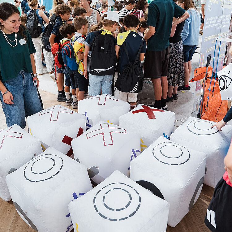 Eine belebte Szene in einer Halle mit einer Gruppe Kinder und Erwachsener, die um mehrere große aufblasbare weiße Würfel mit aufgedruckten Kreisen, Kreuzen und Symbolen stehen; vorne rechts ein Junge im roten Shirt, links eine lächelnde Betreuerin mit grünem Oberteil und Jeans, Rucksäcke bei den Kindern, Holzboden und Informationsstände im Hintergrund.