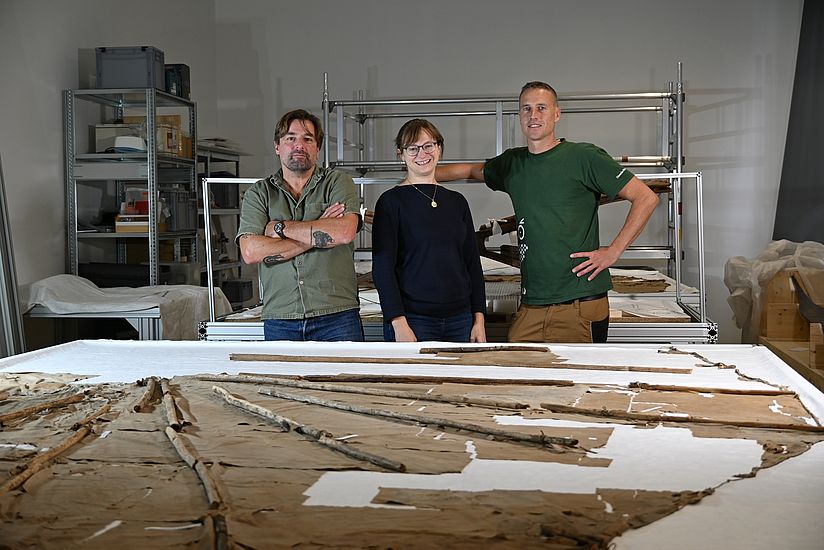 Mathias Winkler, Charlotte Holzer und Patrick Goldbach stehen hinter einem Tisch auf dem der originale Normal-Segelapparat ausgebreitet liegt.
