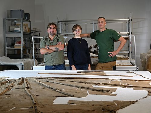 Restaurierungsteam Operation Lilienthal Mathias Winkler, Charlotte Holzer und Patrick Goldbach stehen hinter einem Tisch auf dem der originale Normal-Segelapparat ausgebreitet liegt.