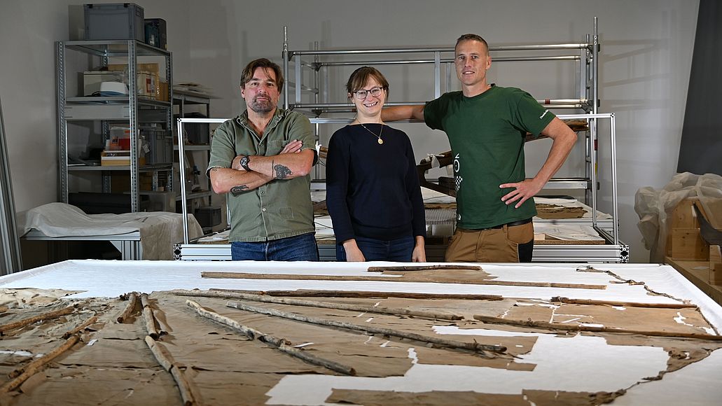 Restaurierungsteam Operation Lilienthal Mathias Winkler, Charlotte Holzer und Patrick Goldbach stehen hinter einem Tisch auf dem der originale Normal-Segelapparat ausgebreitet liegt.