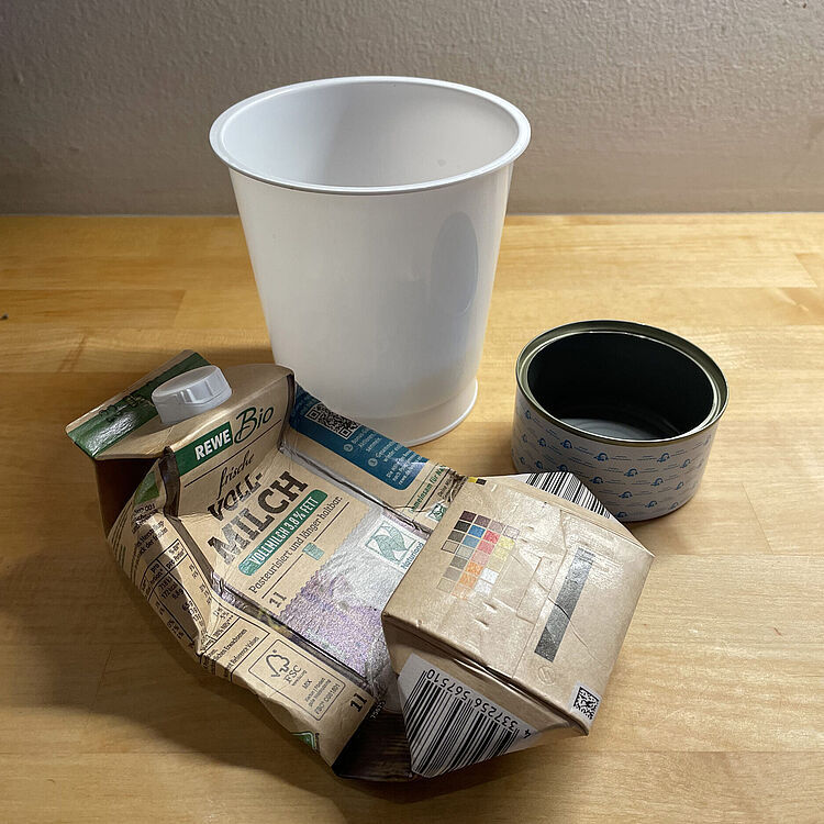 Eine zerknüllte Milchpackung neben einem weißen Kunststoffbecher und einer geöffneten, leeren Konservendose auf einer hellen Holzfläche vor einer hellen Wand.